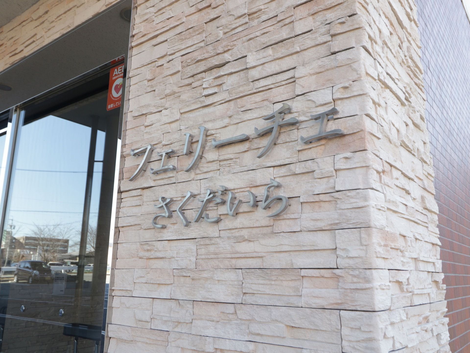 玄関横の白い石造りの壁に、鉄製の部品で施設名が表示されている。ガラス扉にAEDのシールが貼られている。