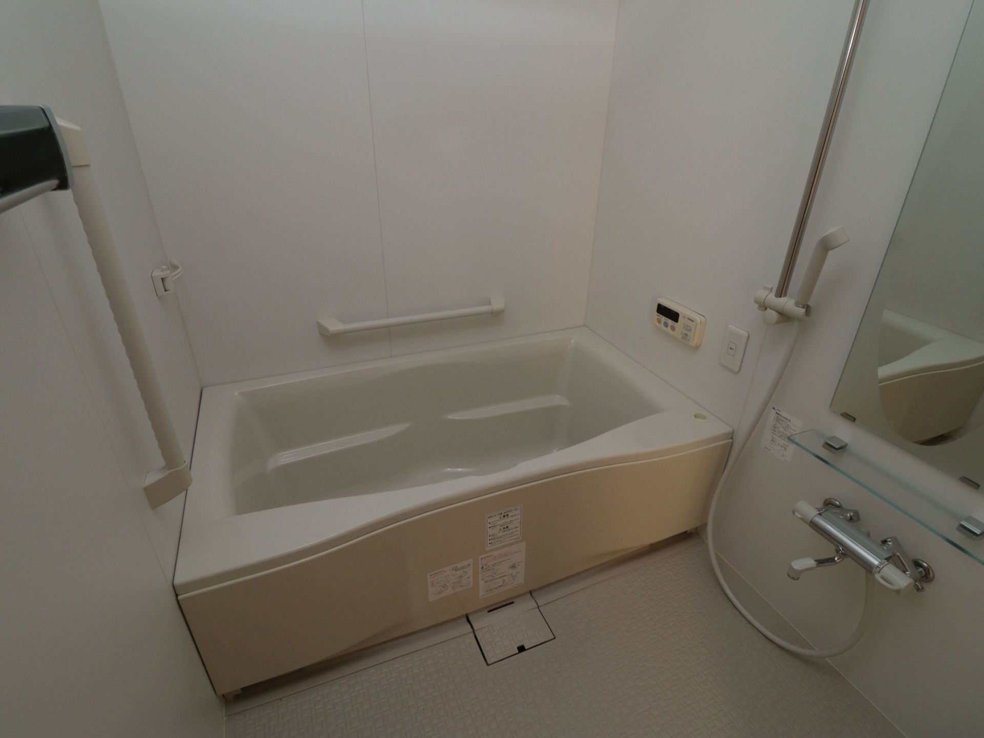白を基調とした個浴用浴室である。壁に給湯用のパネルが取りつけられている。手すりに可動可能なシャワーヘッドがある。