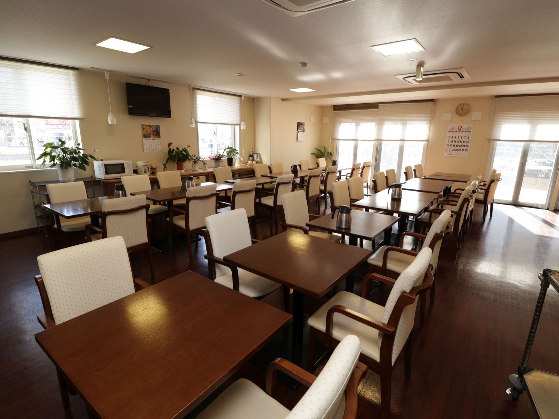 窓がたくさん開いた広間に、２人ずつ利用できるテーブルセットがたくさん並べられている。壁に大型液晶テレビが掛けられている。