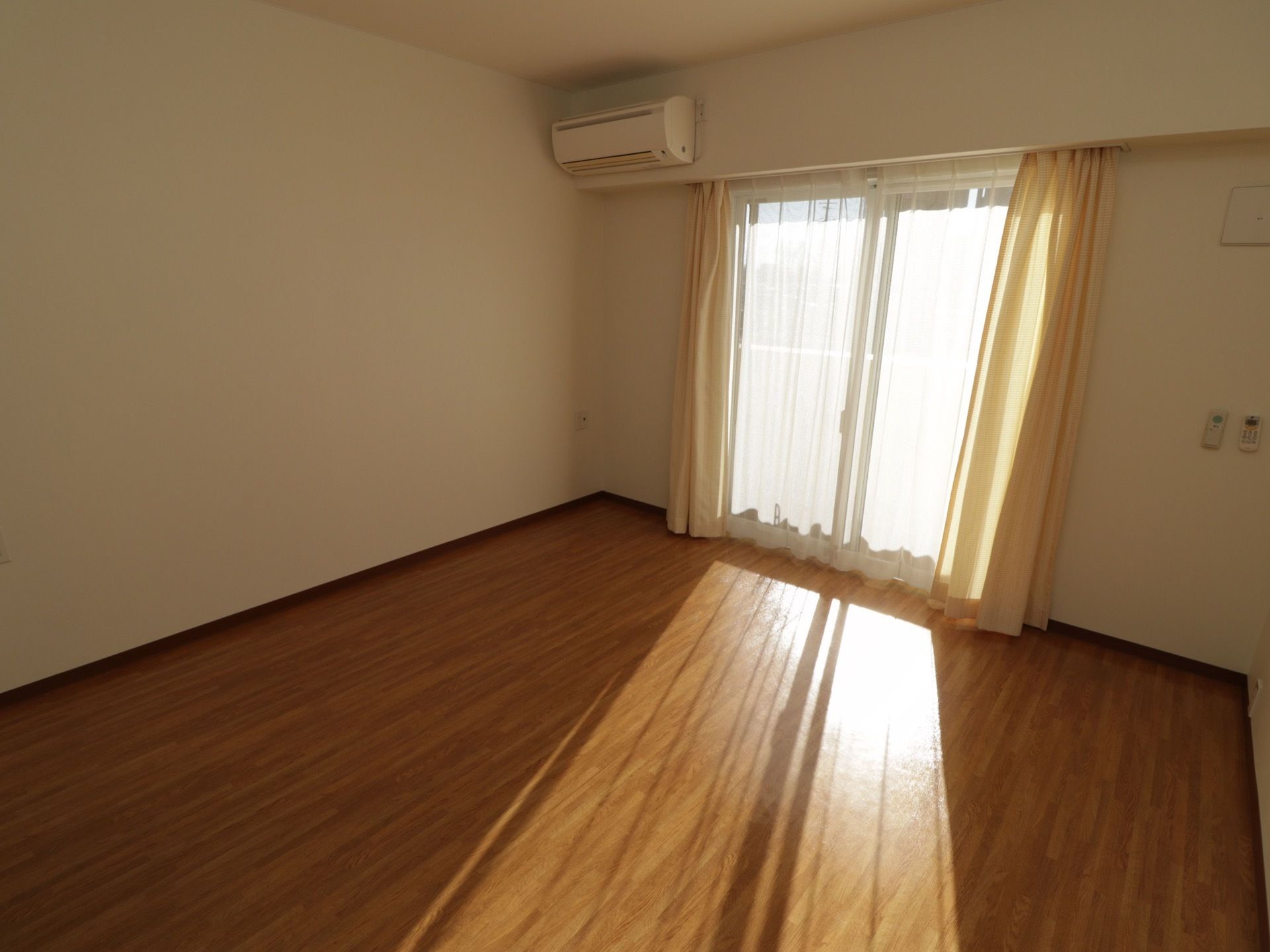 南向きの窓はカーテンがかかっているがたくさんの外光が差し込んでいる。窓の上にはエアコンが設置されている。
