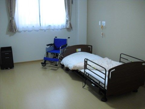 ゆとりある広さのある居室の中に、介護用ベッドと車椅子、チェストが置かれている。部屋の奥にある窓から明るい光が射し込んでいる。