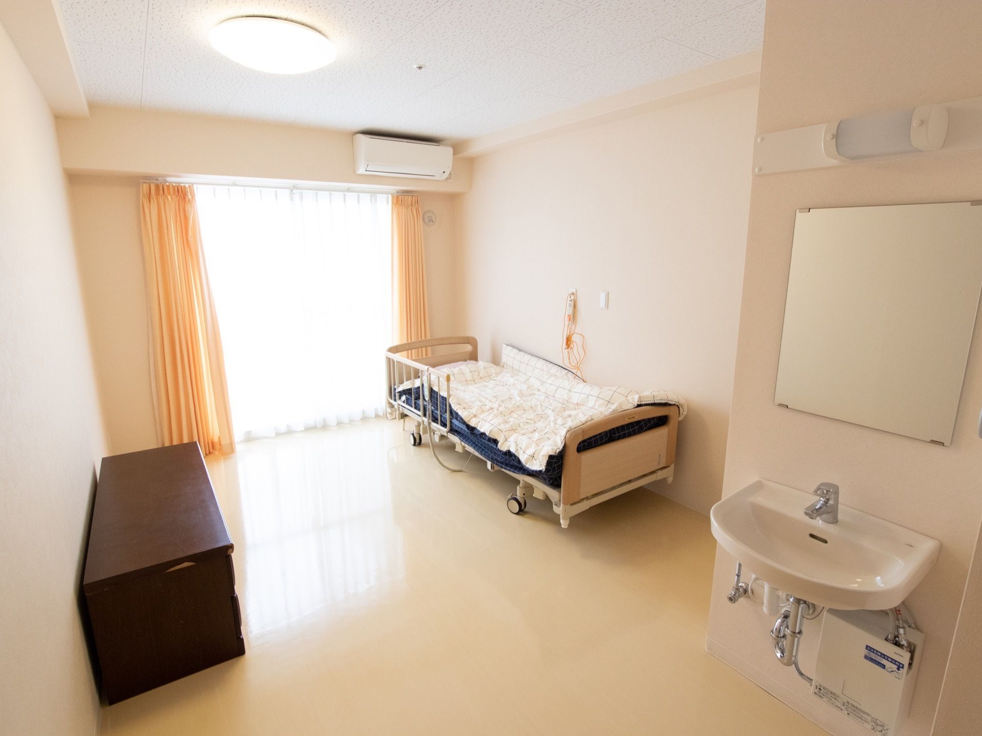 白を基調としたデザインの居室である。介護用ベッドとタンスが置かれている。壁には、洗面台と緊急時用のコールが設置されている。