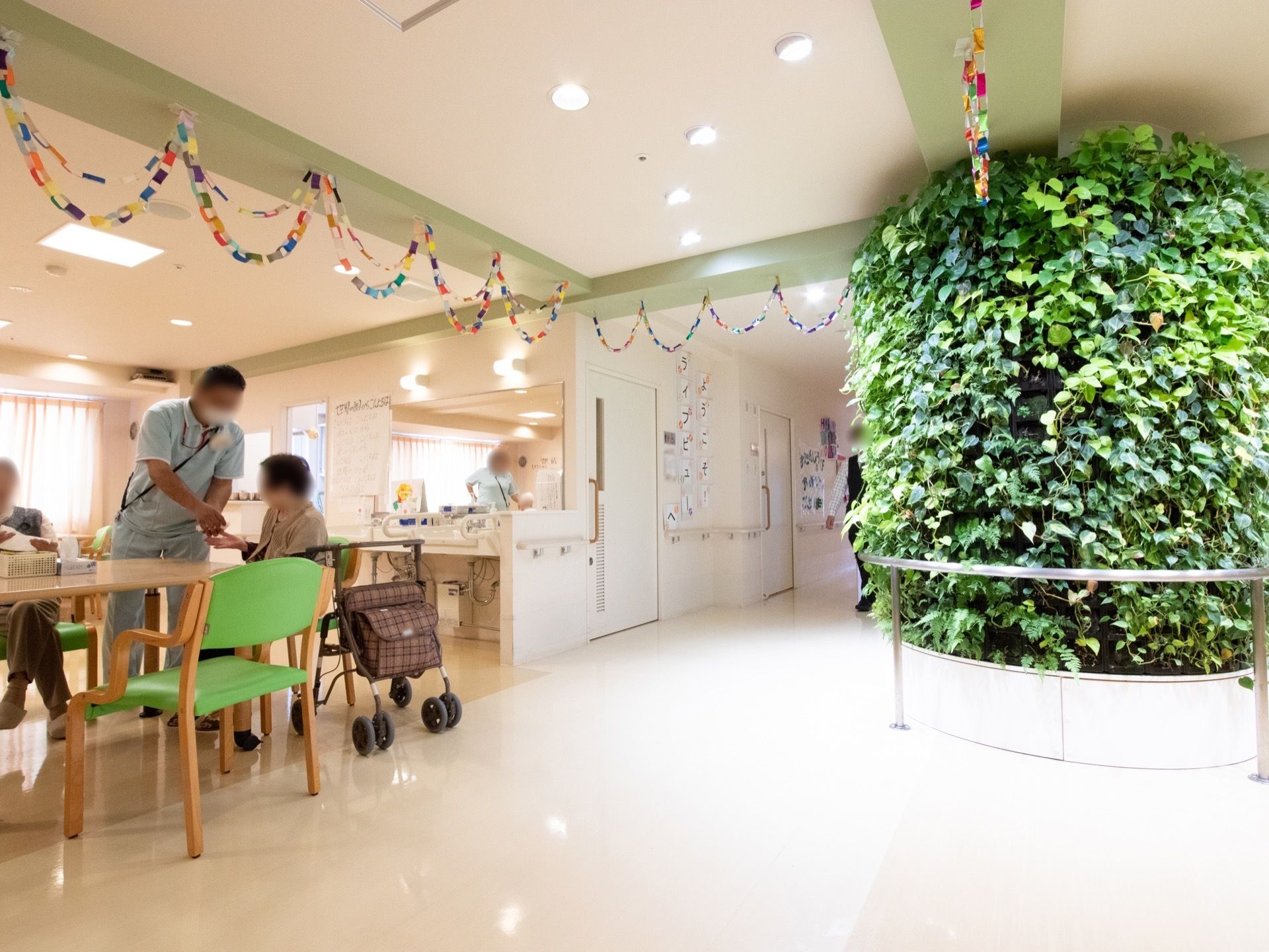 エントランスの中央の柱は、一面が植物で覆われている。広々とした共有スペースがあり、机や椅子が置かれ、職員とレクリエーションを行っている。