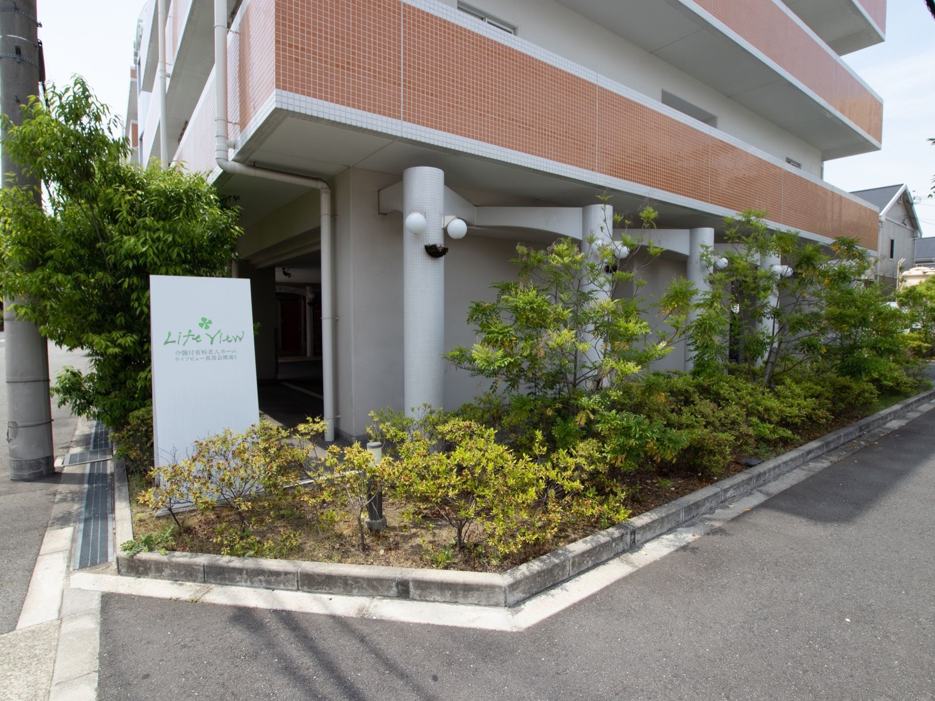 施設の建物は、道路に面する。洋風な外灯があり、バルコニーの外壁はタイル張りである。植え込みに樹木が植えられている。