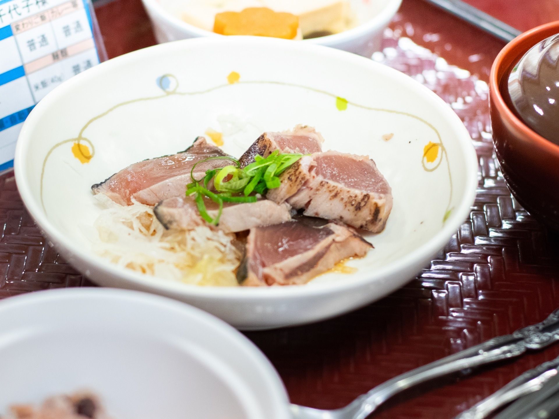 お盆の上に和の献立と食器が並んでいる。主菜は、大根のつまの上にカツオのたたきがのっている。ご飯茶碗に椀物、小鉢には煮物が入る。