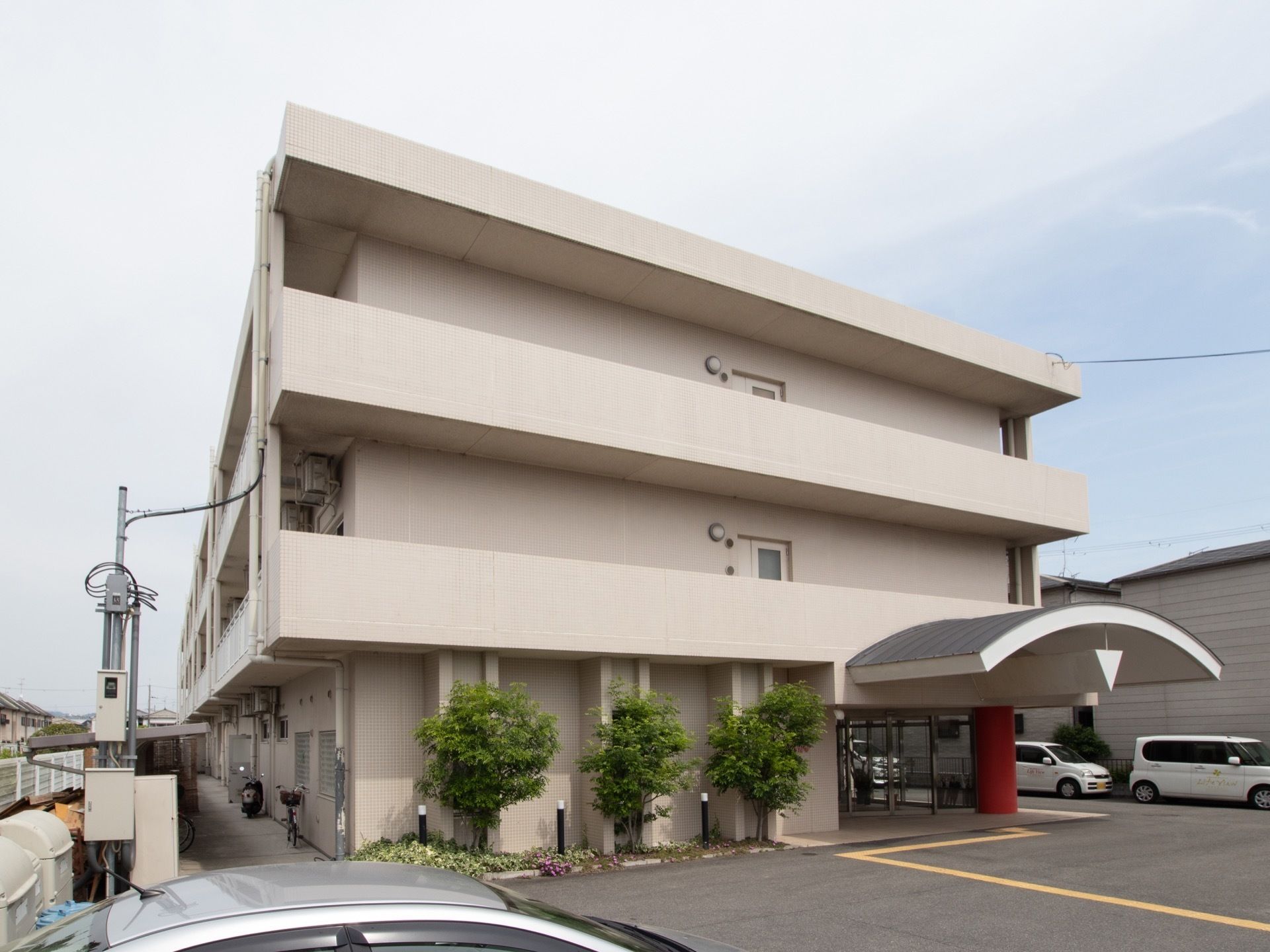 三階建ての薄い桃色をした建物。入り口付近には、緑色の木や花のついた茂みなどがある。また、広い駐車場がある。