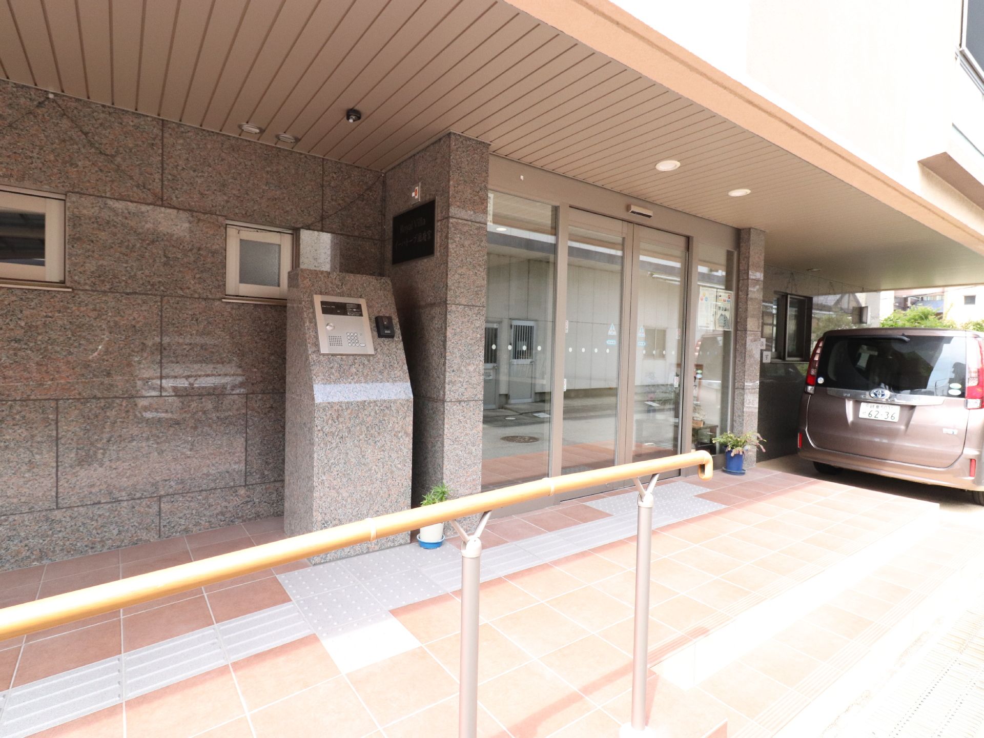 施設の玄関前には、なだらかなスロープがあり、手すりが取り付けてある。駐車している車がそばにある。プランターが置いてあるのが見える。