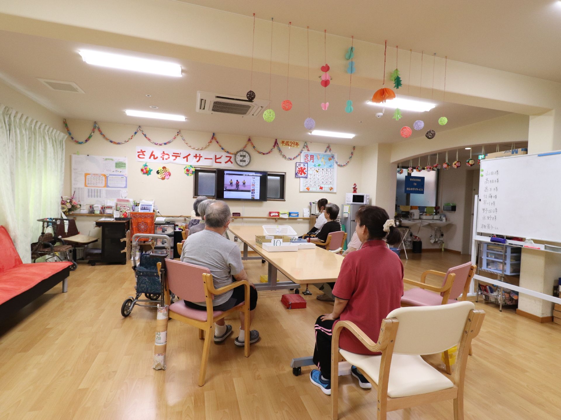 食堂としても利用している部屋は、多目的に利用され、今は職員と入居者がテレビを見ている。天井から手作りのガーランドが釣り下がっている。