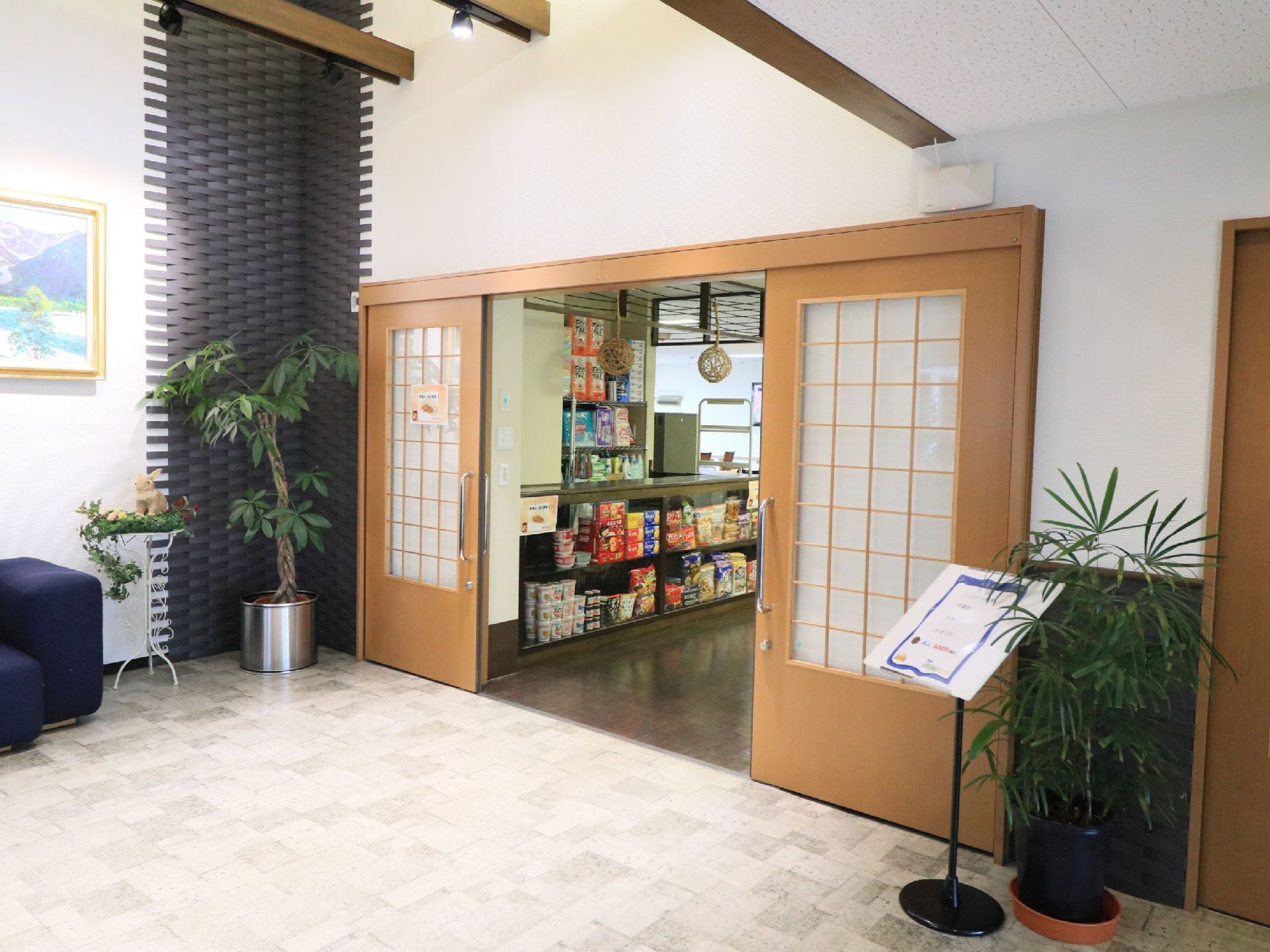 格子状のデザインの扉が設置されていて、扉の横には観葉植物などが置かれている。扉の向こうに売店があるのが見える。