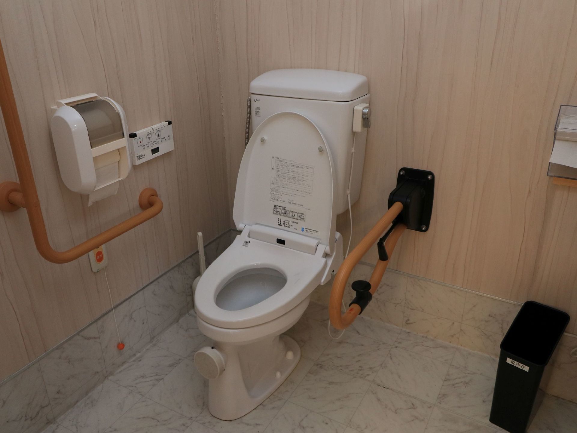 白い大理石調の床に白いトイレが設置されている。トイレの横には可動式の手すりとＬ字型の手すりが取りつけられている。