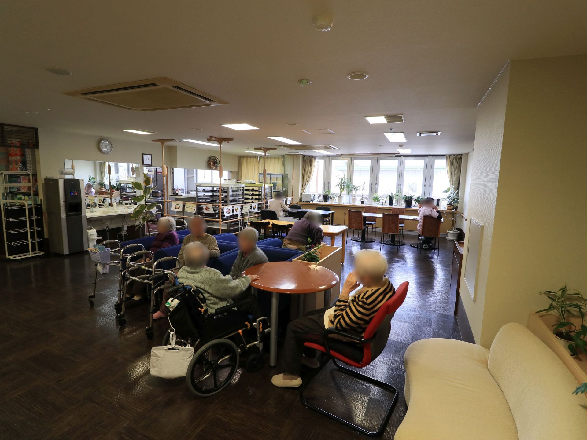 青い１人掛けのソファや円形のテーブル、白いソファなどが設置されていて、入居者様数人が座っている。横に観葉植物が置かれている。
