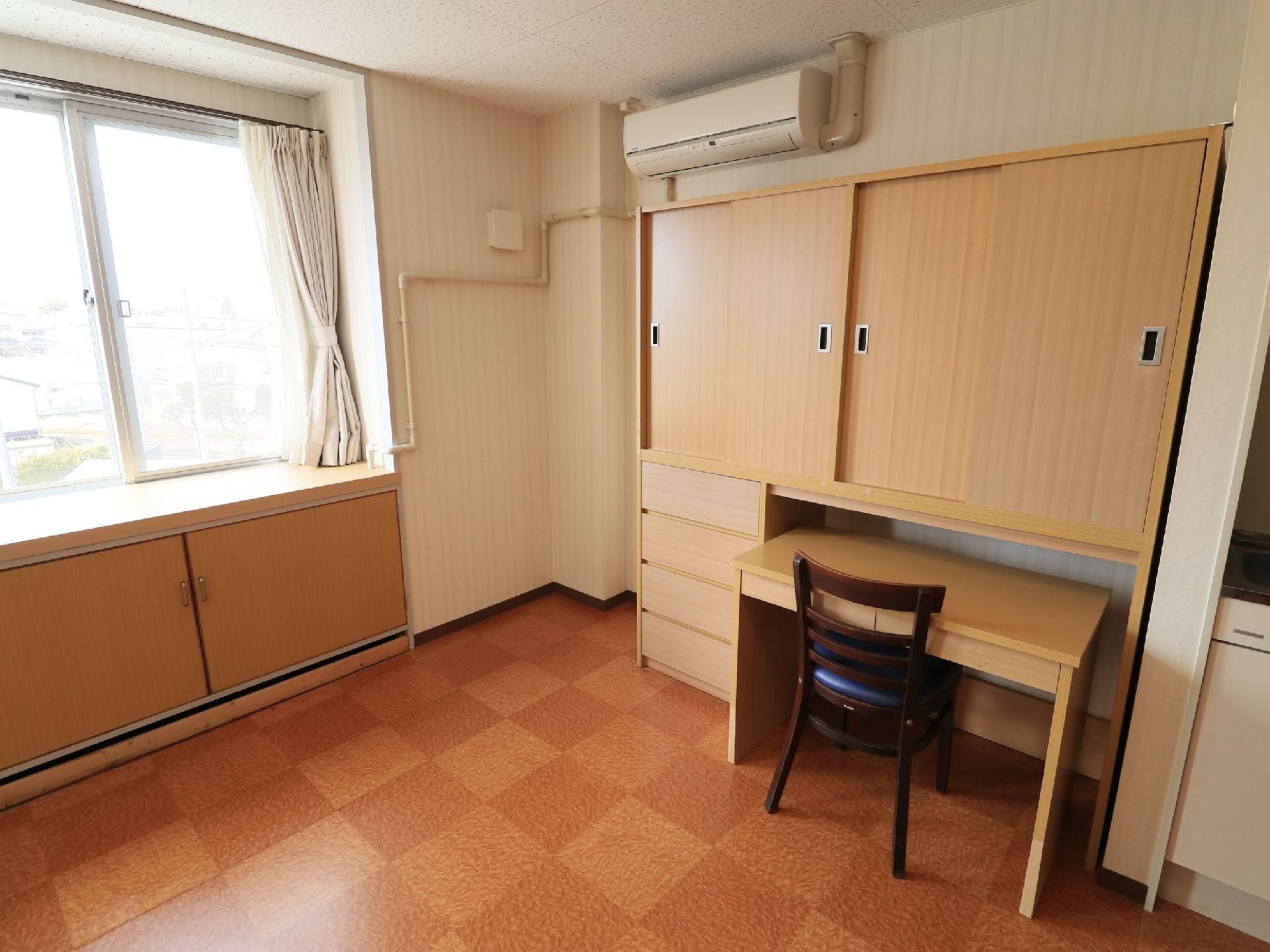 居室内の壁にはエアコンが取りつけられていて、その下に木目調の収納スペースとデスク、椅子が設置されている。