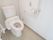 白でまとめられたトイレである。壁にはL字型の手すり、緊急時コールボタンが設置されている。温水洗浄便座もついている。