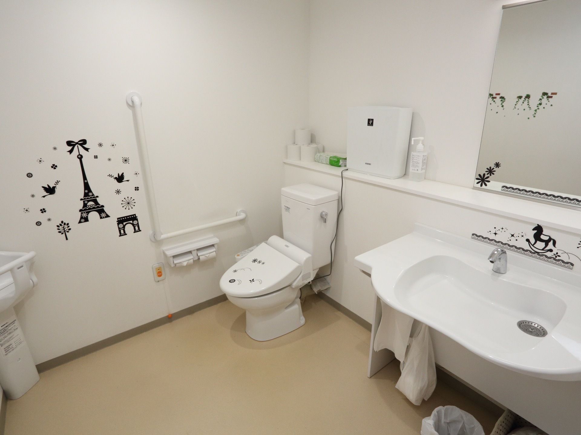 広い共有のトイレがある。白で統一された清潔な空間となっている。洗面台と空気清浄機がある。緊急用ボタンが設置されている。