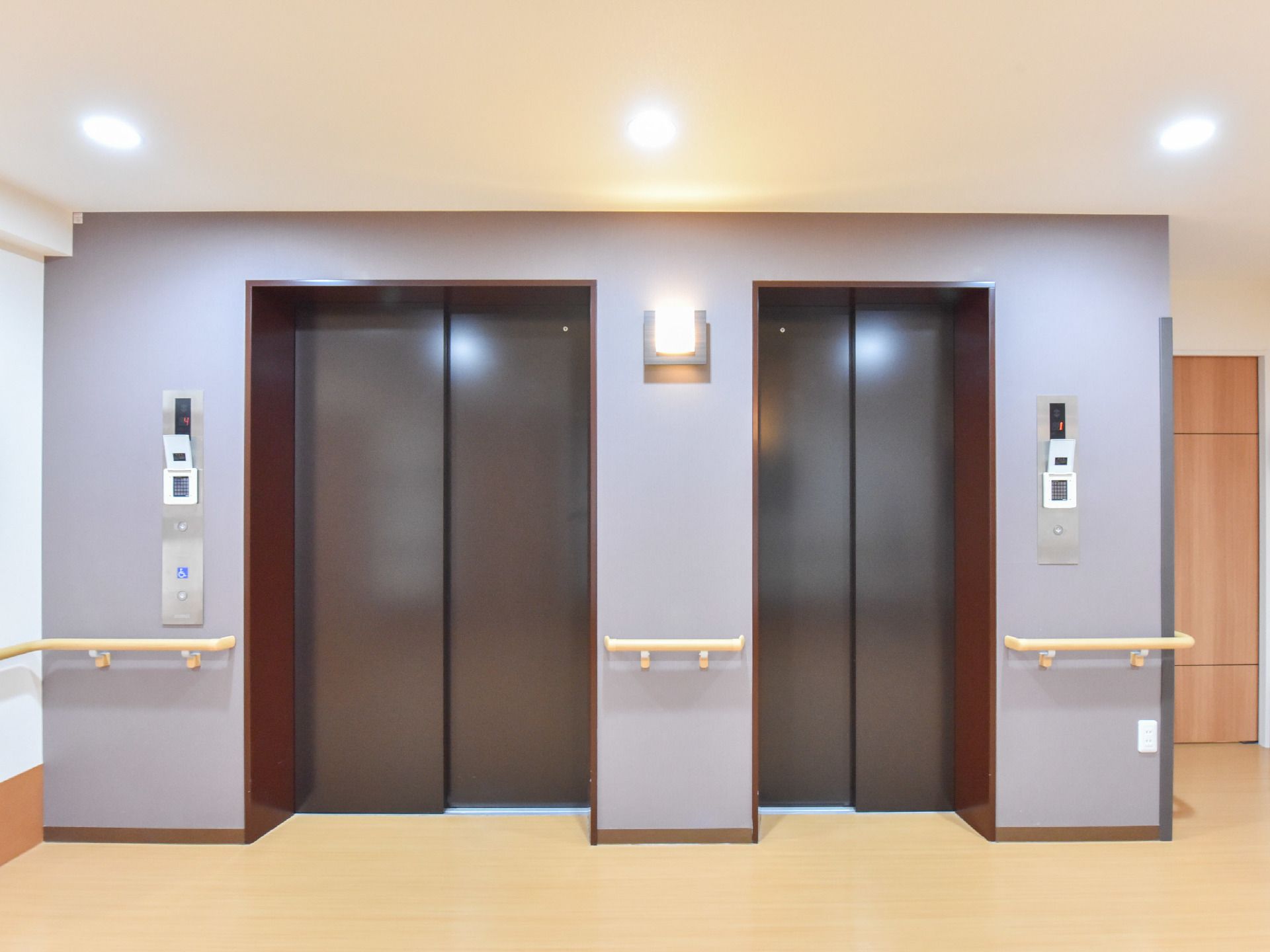 こげ茶色のドアが付いた2台のエレベーターが並んでいる。それぞれ大きさが異なっている。壁にはベージュ色の手すりもある。