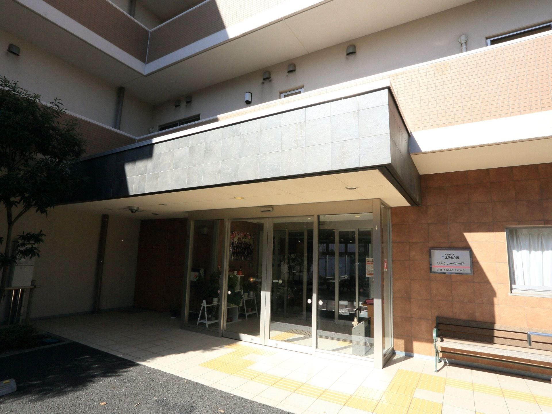 ガラス張りの自動ドアの上に、大きなひさしが設置されている。黄色い点字ブロックが、周辺の地面に埋め込まれている。