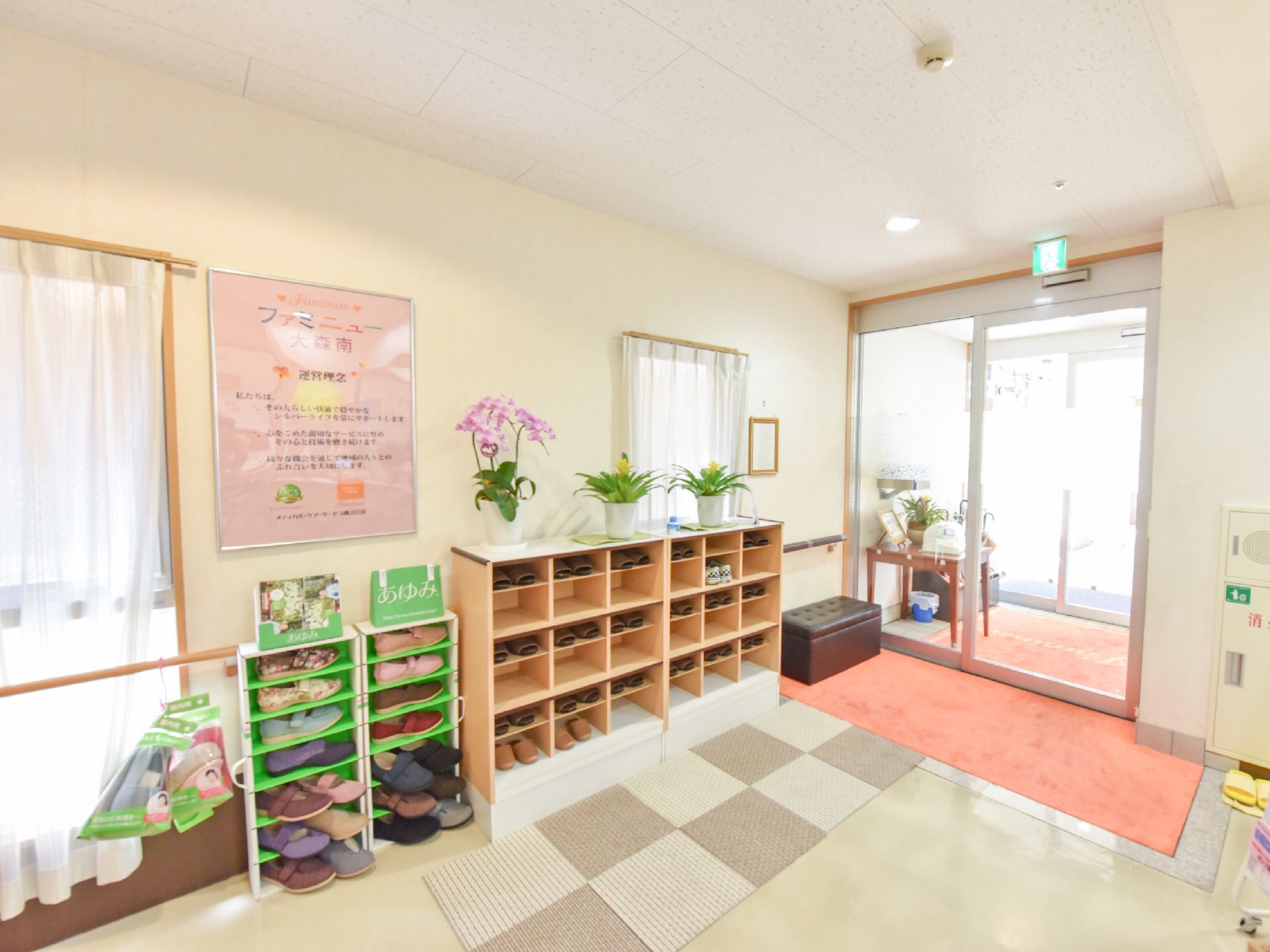 下駄箱の上に花の鉢が有り、その隣にはスリッパが入った棚がある。窓が幾つかあり、明るい光が差し込んでくる玄関ホール。