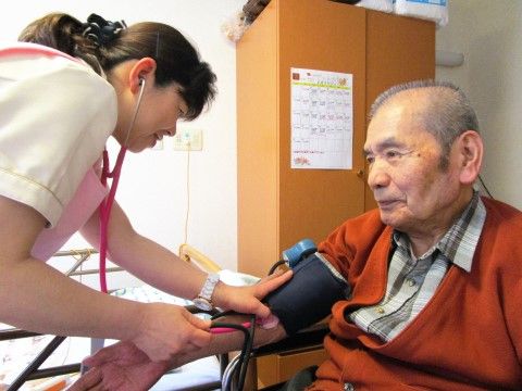 笑顔で血圧を測る介護スタッフと、椅子に腰かける高齢男性