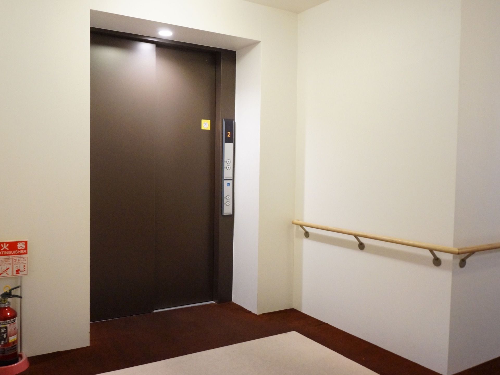 エレベーターの周囲の壁は白く、ナチュラル系の色の手すりが取り付けられている。エレベーターのそばには、消火器が設置されている。