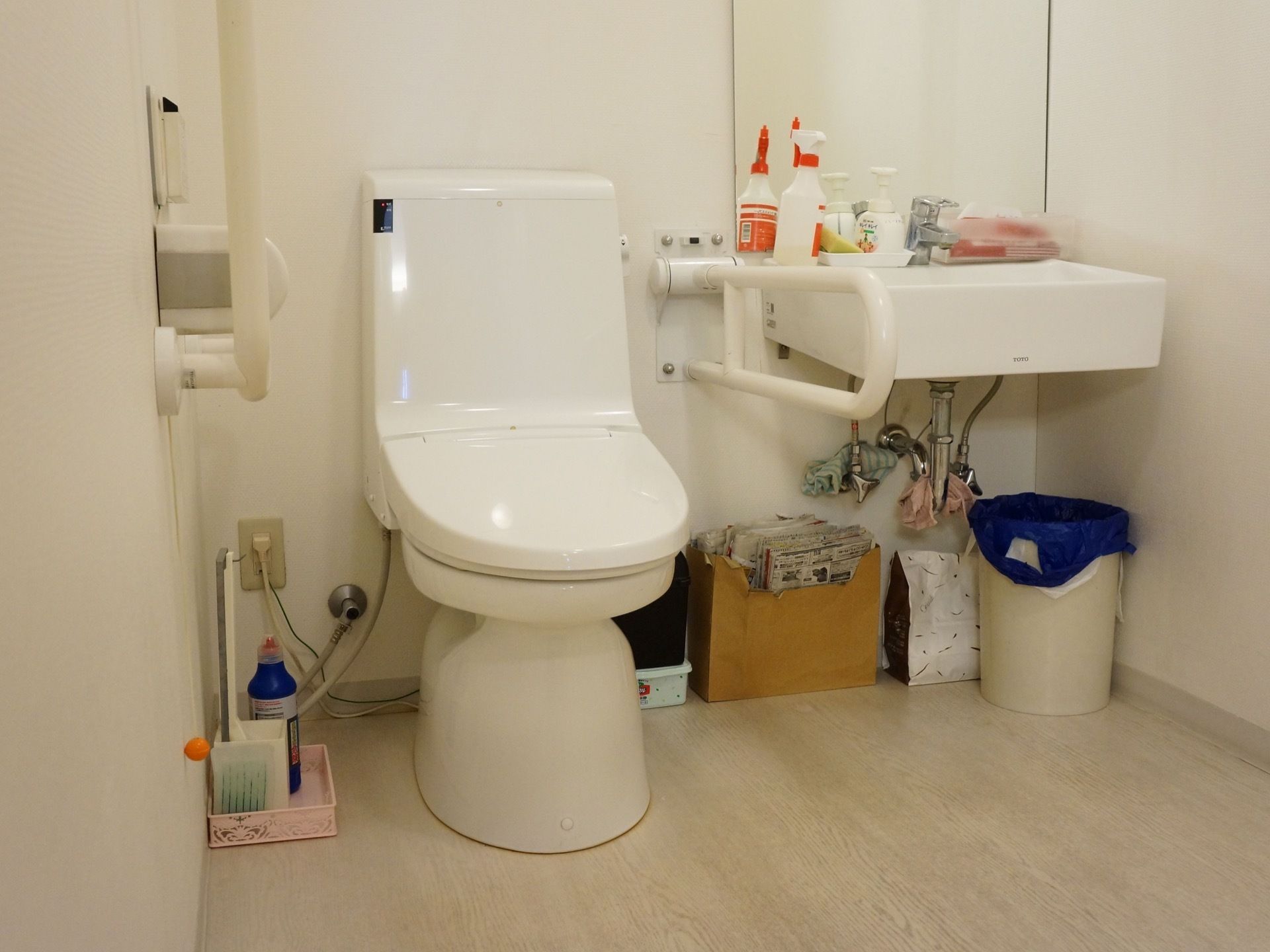 トイレは洋式である。便器の両側に手すりがあり、入居者様が姿勢を楽しむのに役立っている。洗面台の周囲には清掃道具がある。