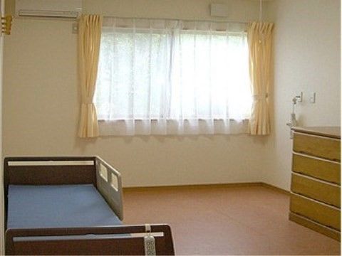 右手に木製のチェスト、左手にガード付きベッドが設置され、オレンジ色のカーテンのかかった窓からは明るい日が差し込む個室