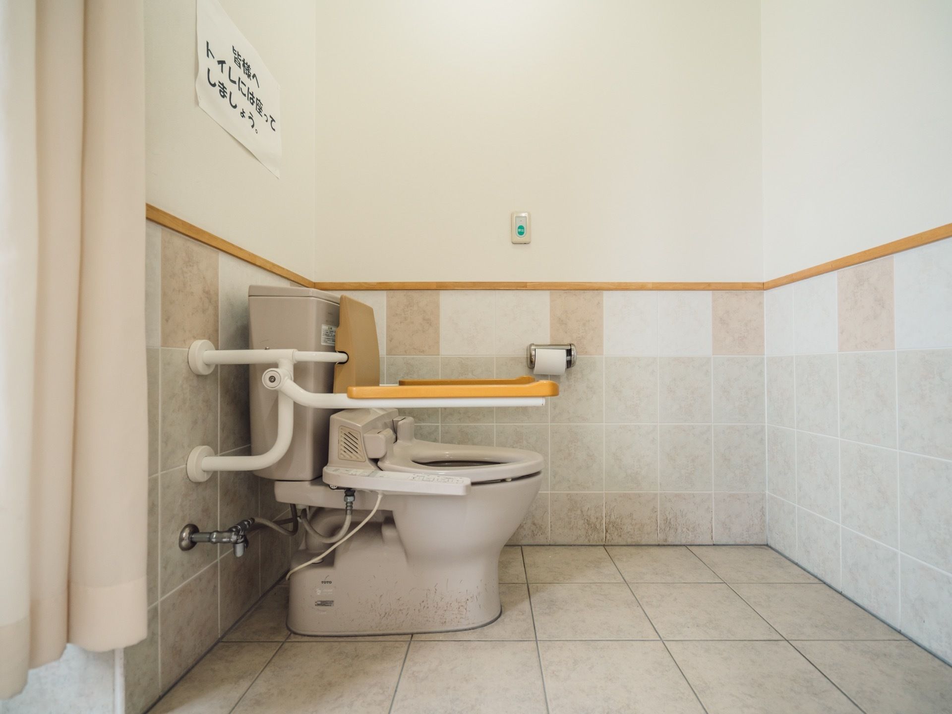 トイレは広く、タイル張りの床で統一されている。入口にはカーテンが掛けられており、手すりや背もたれも取りつけられている。