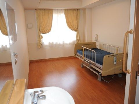 カーテンが半分開かれ、高級感がある居室になっている。介護ベッドにはサイドレールが設置され、リモコンが掛けられている。