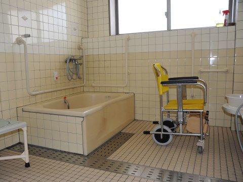 タイル張りの浴室である。中央に排水溝が並んでおり、金属の蓋がされている。角に浴槽があり、洗い場には車椅子タイプのバスチェアがある。