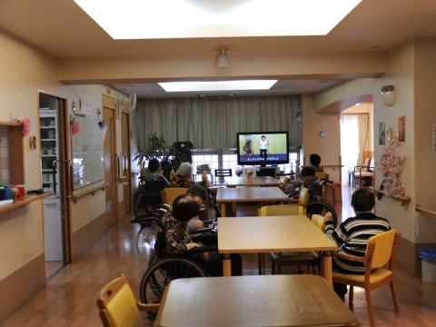 木製テーブルを囲むように座りながら液晶テレビを眺めて過ごす人々がいる広間