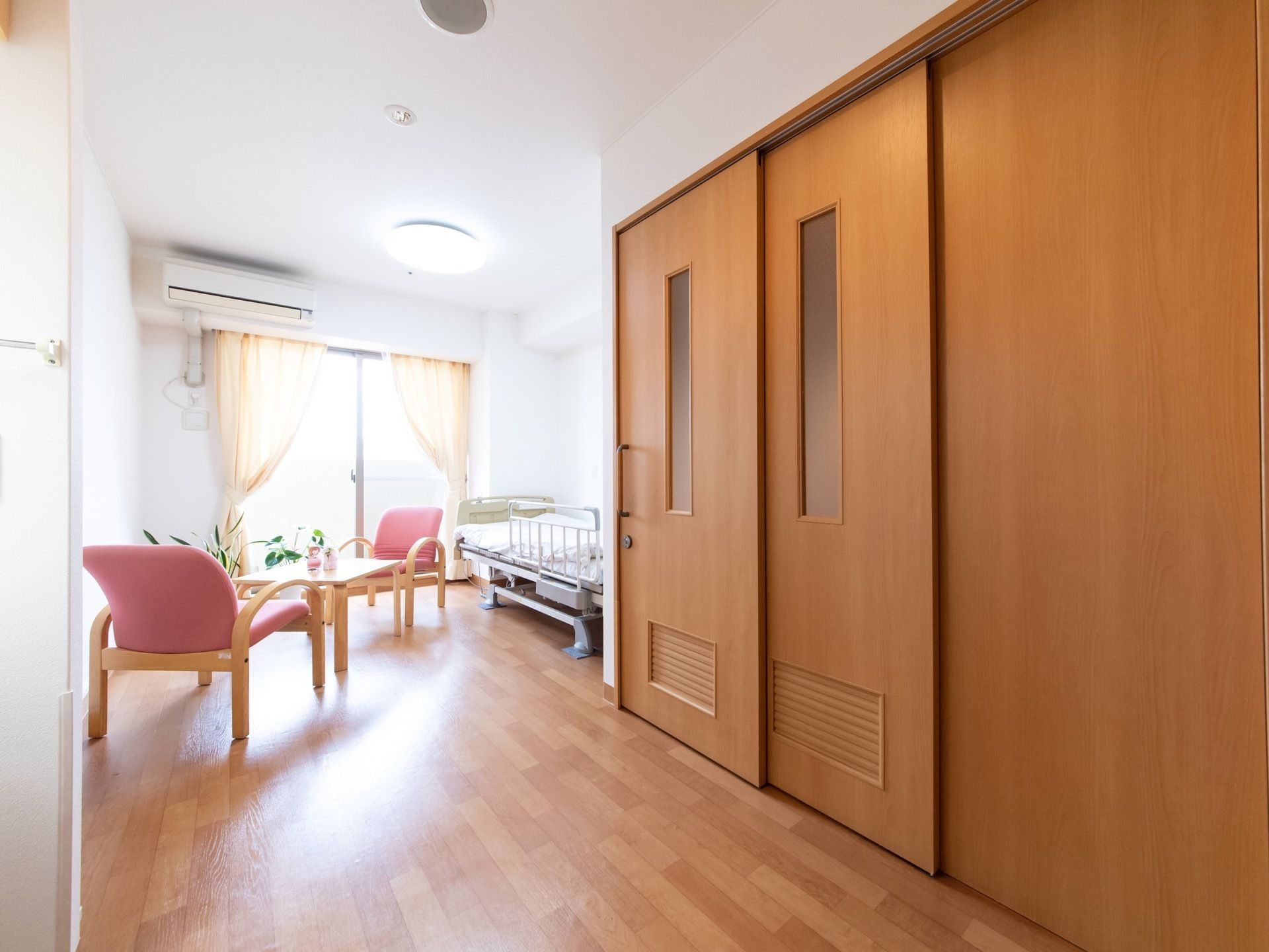 居室の窓辺にベッドが置かれており、朝は自然光で健康的に目覚めることができる。テーブルセットと観葉植物が設けられている。