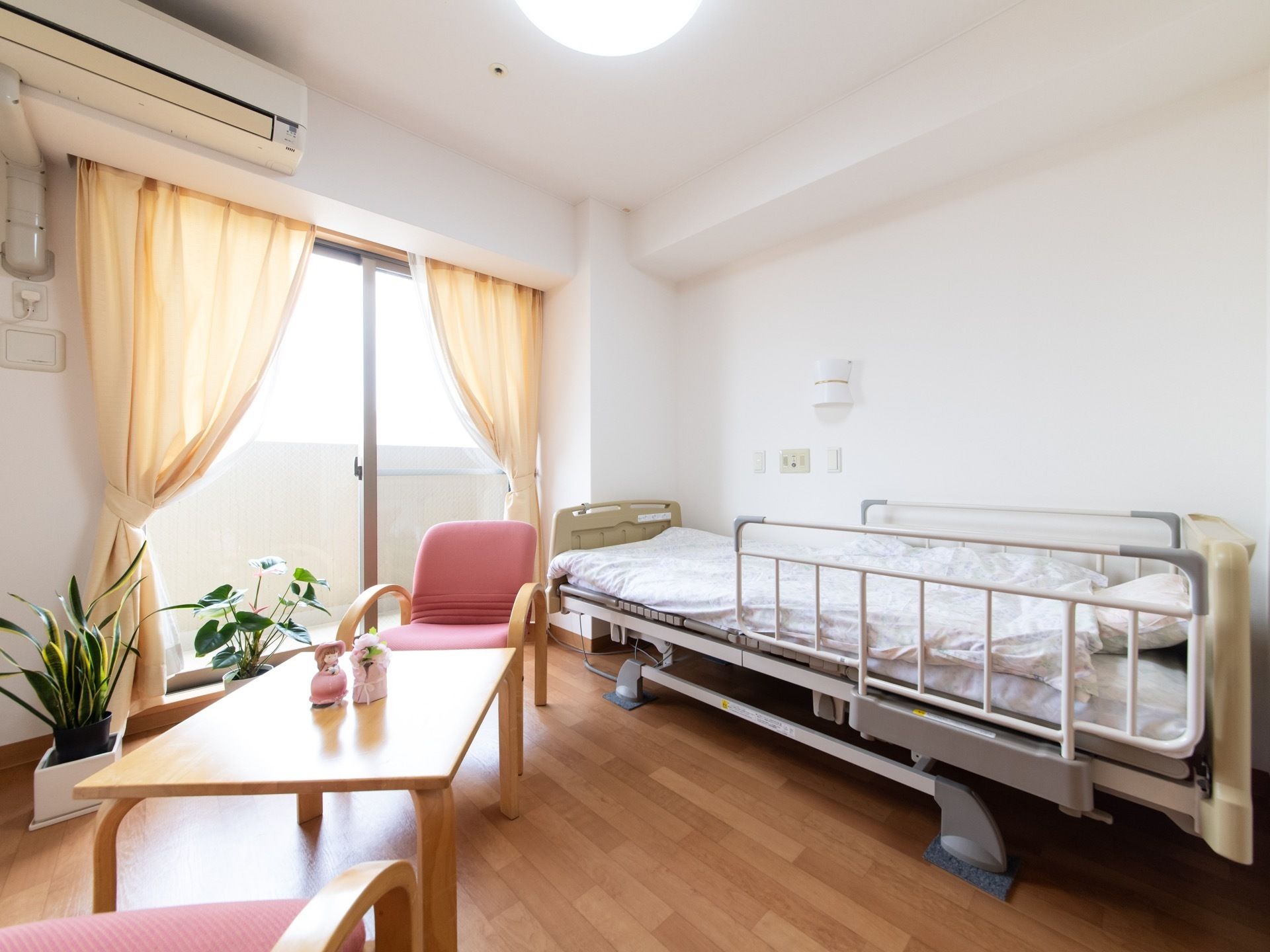 居室には電動ベッド、2脚の椅子が付いたテーブルセット、エアコン、照明が備わっている。白壁の部屋に緑の観葉植物がワンポイントとなっている。