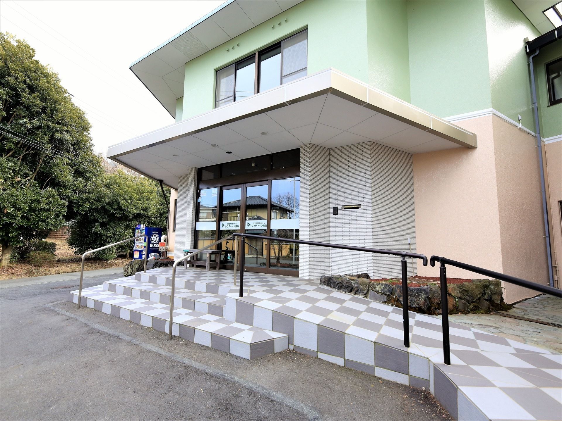 玄関ポーチはグレーと白の市松模様のタイル敷きである。出入り口の右側にスロープと手すりが取りつけられている。