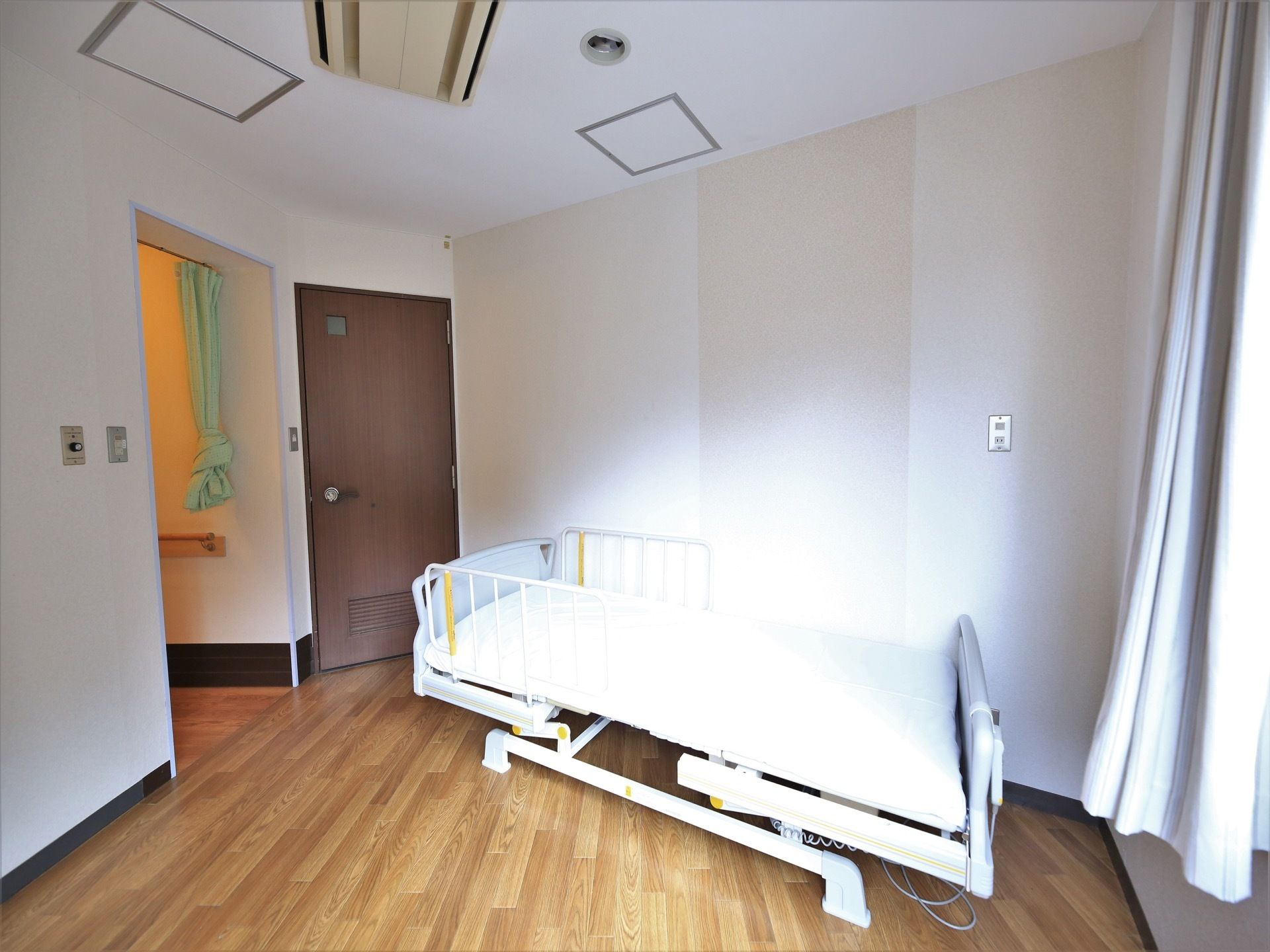 フローリングの床の居室内にベッドが置かれている。茶色いドアがあり、その横にはグリーンのカーテンが掛かっている。