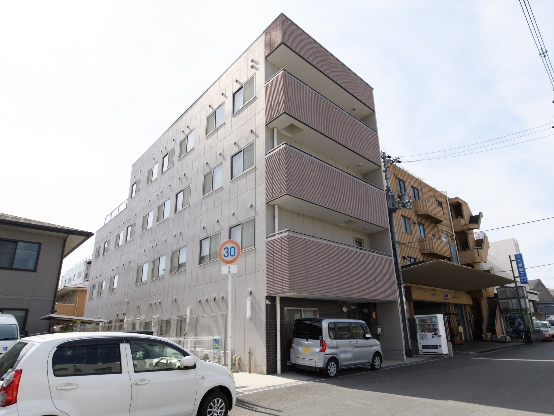 ４階建てのコンクリート調の各階にベランダがついた建物である。建物前の道幅が広く軽自動車が１台停まっている。