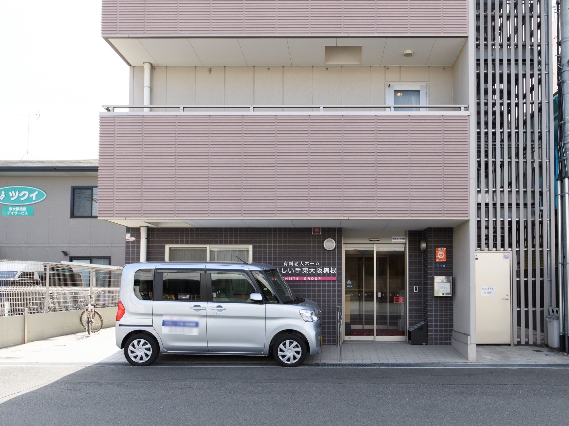 １階のタイル調の壁に施設名が表示されている。自動ドアの手前に傘立てが置いてある。敷地内に軽自動車が１台停まっている。
