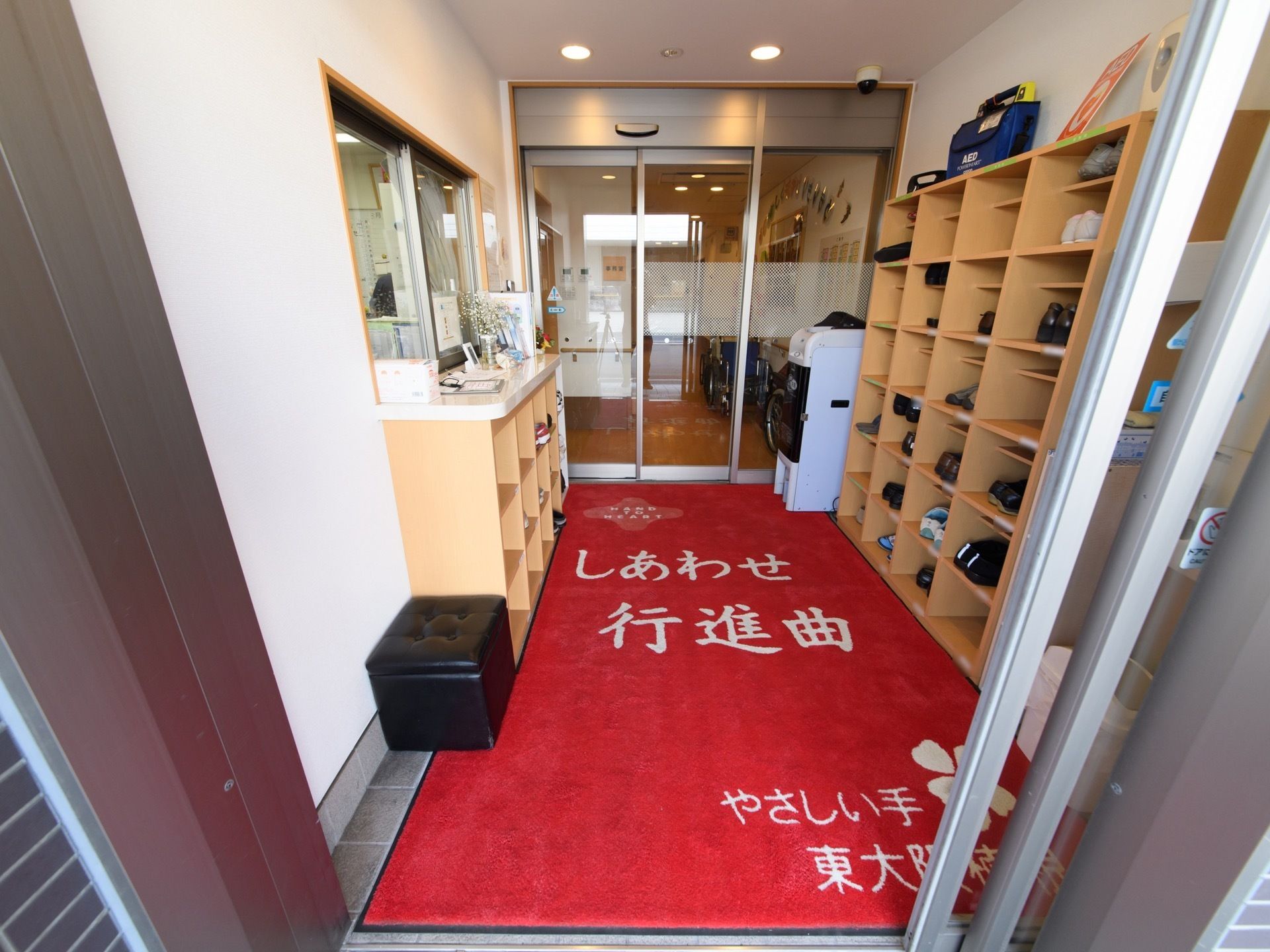 通路の両側に個別に入れられる靴箱が設置されている。小さな黒い腰掛が１台用意されている。自動ドアはガラス張りで室内の見通しが良い。