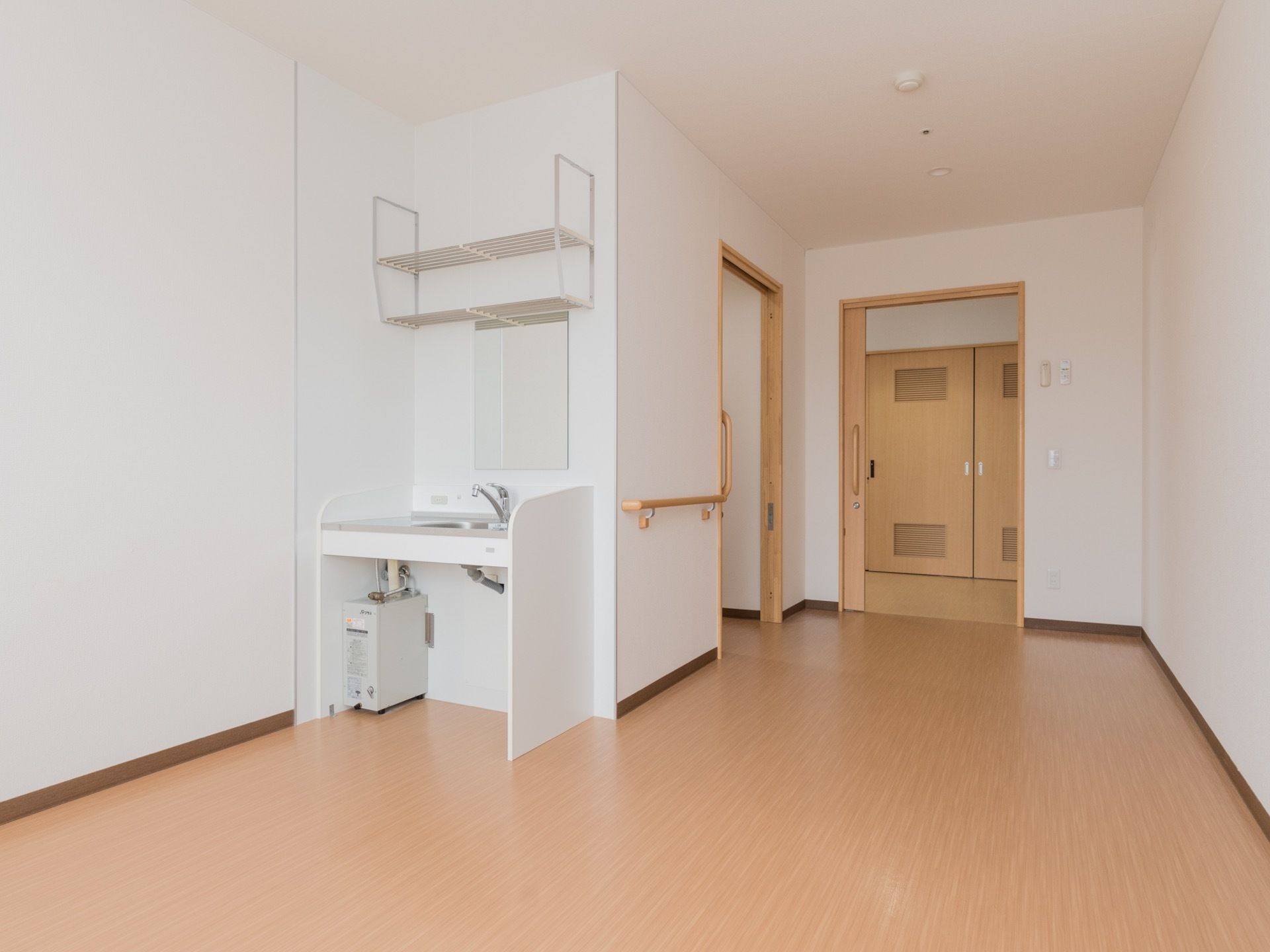 居室の扉は開けられている。中は広々としている。壁の一部には手すりが取り付けられている。鏡付きの洗面台の上には棚がある。