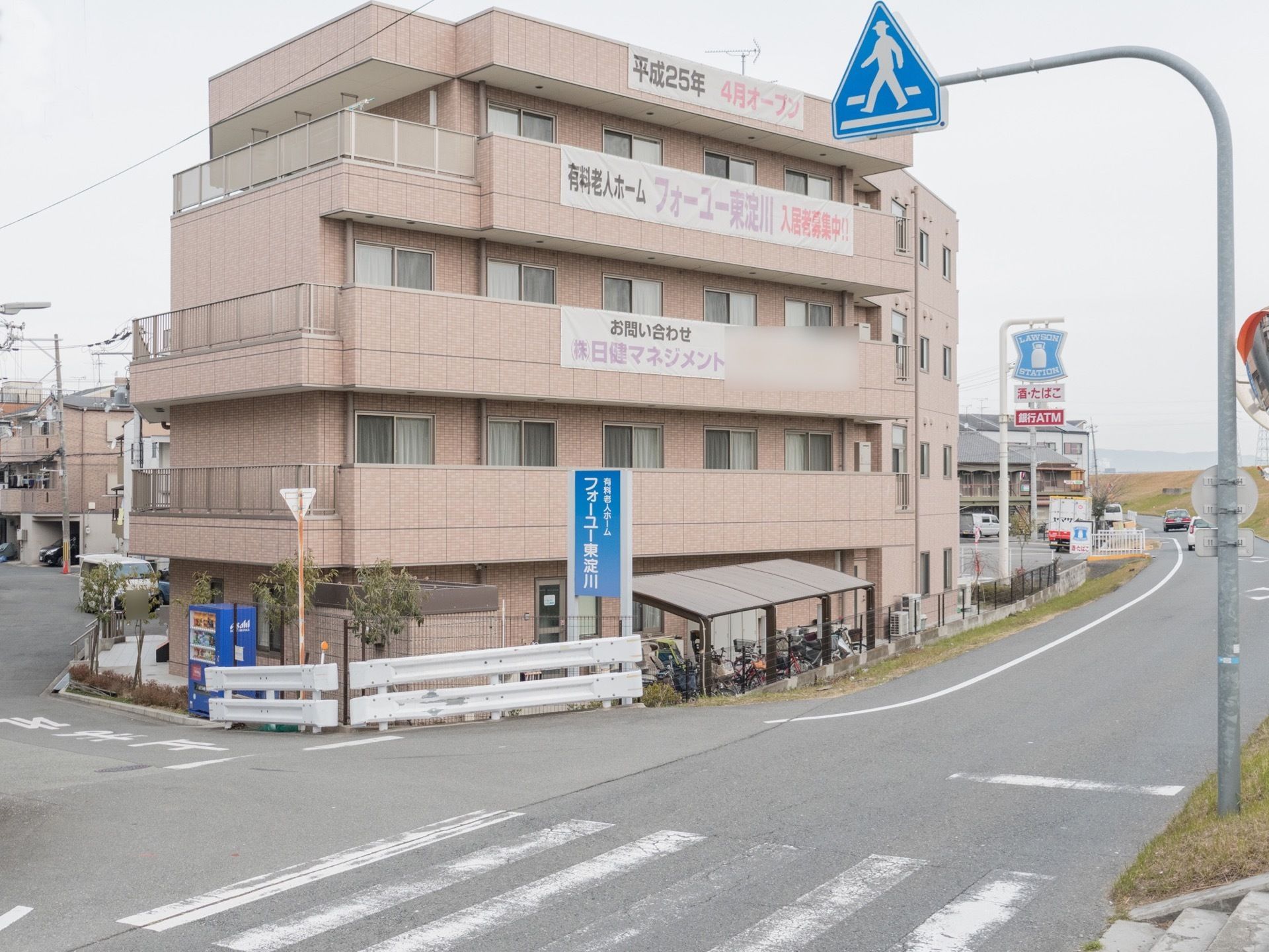 施設は道路に面して建てられた4階建てである。周りは緑に囲まれている。外壁には入居者様募集の幕がかけられている。