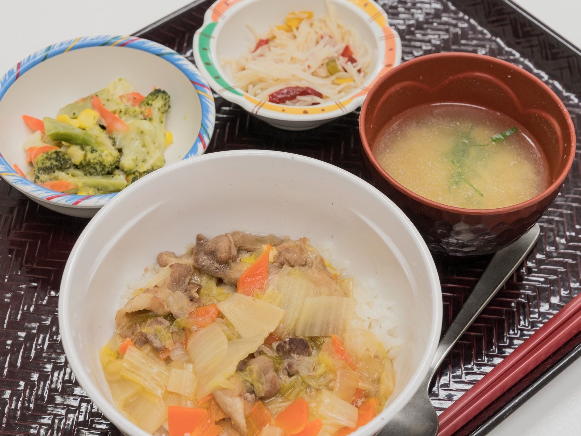 黒っぽいお盆の上にはその日の食事が配膳されている。メインの中華丼をはじめ、ブロッコリーと人参、コーンのサラダなどの4品がある。