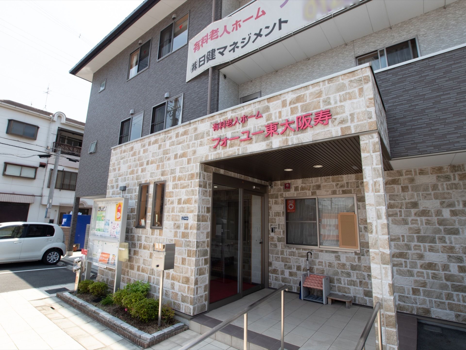 玄関前に大きな掲示板があり、その横にポストがある。入口前に傘立てが置かれている。屋根に施設名が表示されている。