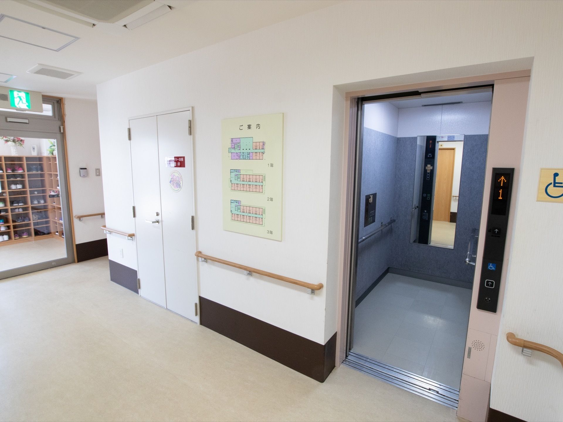エレベーターの扉横には、館内案内図が貼られている。壁には手すりが取り付けられ、奥は玄関になっている。