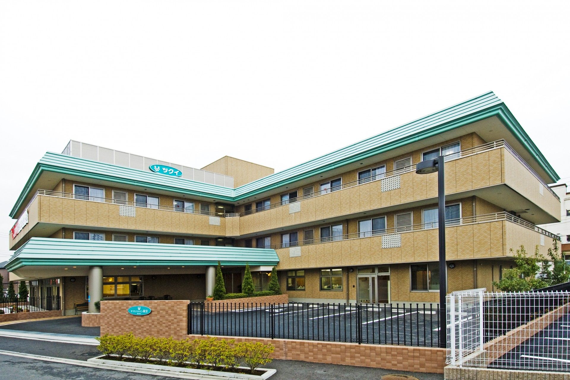 ブラウンの外観にミントグリーンの色の屋根がついている。駐車場が設けられている。周囲はフェンスで囲われている。