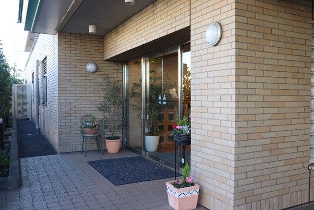 玄関はガラスドアで明るい。玄関前には椅子を置いた喫煙コーナーと傘立てがある。1階駐車場から大きな庇が続いており、雨の日も濡れずに入れる。