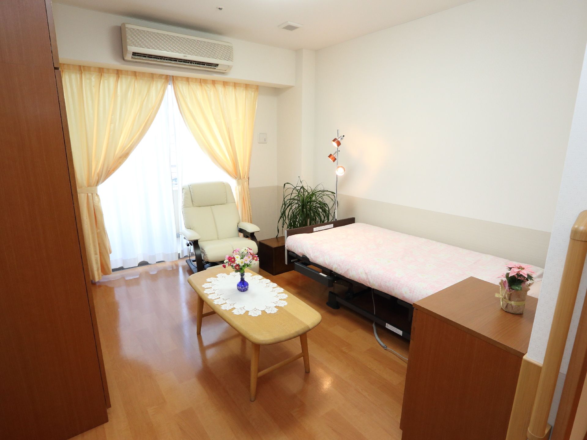 リモコンのある部屋で、窓には黄色のカーテンが取り付けてある。左手に収納棚がある。テーブルの上には、花が飾ってある。