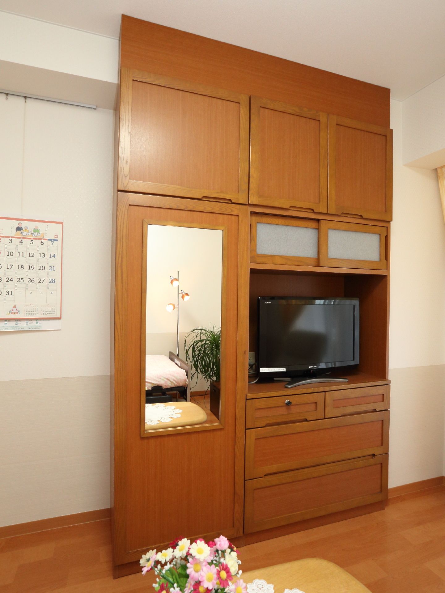 茶色のフローリングの床の上に、茶色の木製のテレビラックがおいてあり、テレビが設置されている。壁にカレンダーがかけてある。