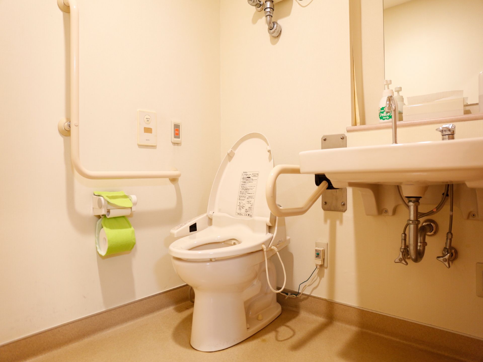 洋式と入れがあり、手すりが壁に設置してある。黄色のカバーがかかったトイレットペーパーホルダーがある。