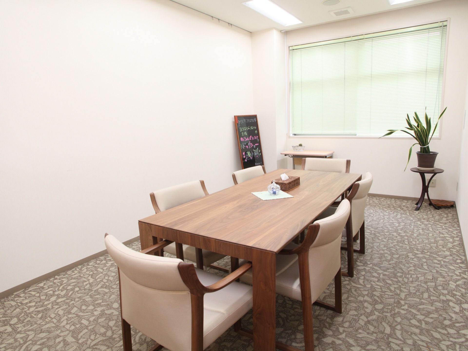 縦長のテーブルに６脚の椅子をセットした部屋は、面接・相談などに用いている。茶系の家具と植物やサンシェイドの緑色の色合いがマッチしている。
