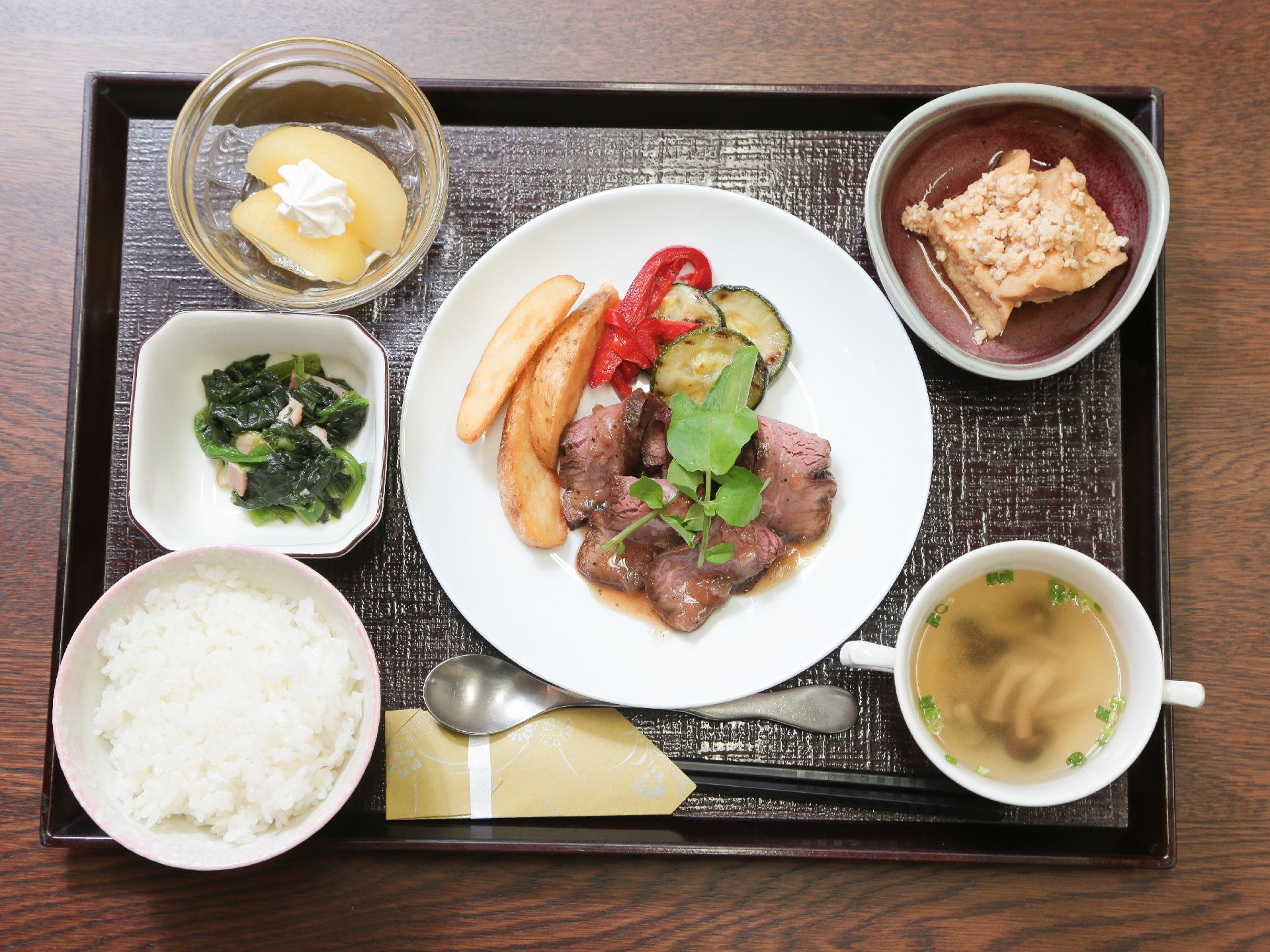 トレーの上にご飯と汁物、真ん中にメインおかずがあり、小鉢が3つ添えて置かれている。スプーンとお箸がある。