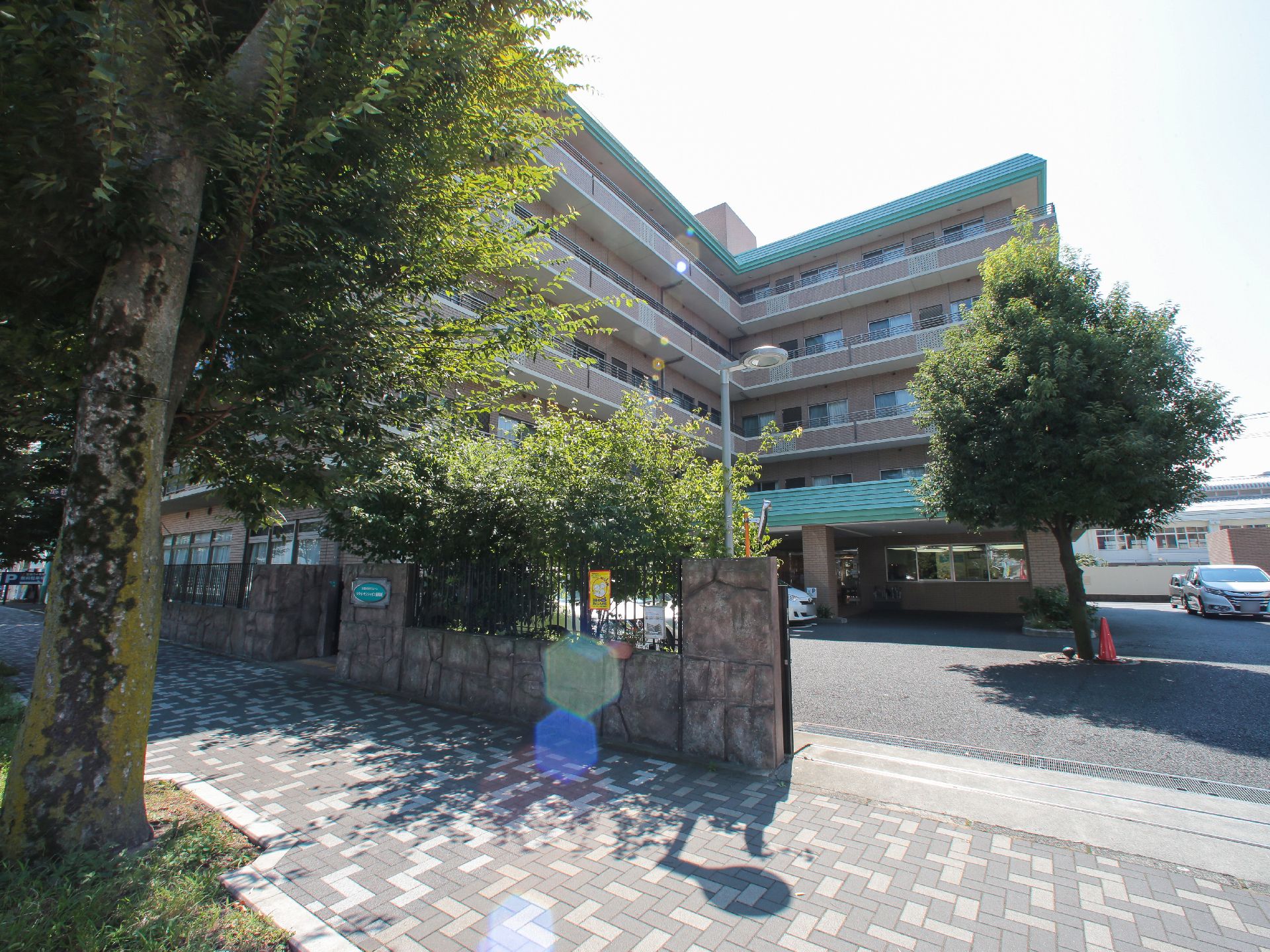 グレイのツートンカラーのタイル調の歩道沿いに面した施設である。薄い茶色と薄い緑色の外観の5階建ての建物である。
