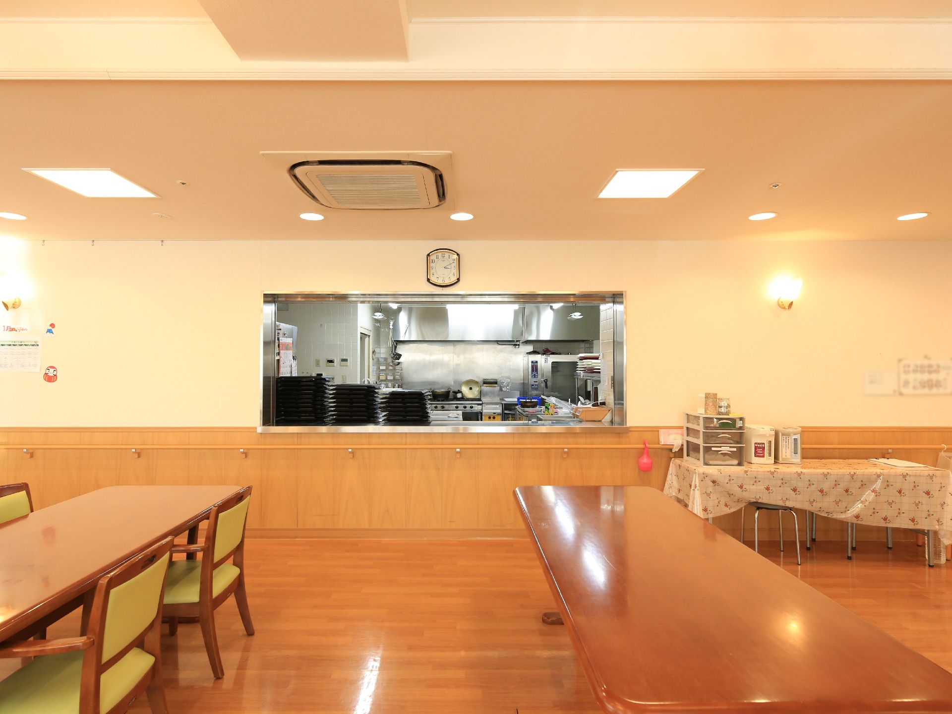 食堂の一角にある、厨房へと続くカウンター。テーブルと椅子が置かれ、床と壁の下部分は木のぬくもりが感じられる素材でできている。